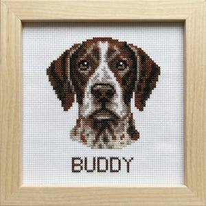 May include: Cross-stitch portrait of a dog with brown and white markings, framed in light wood. The dog's name, "BUDDY," is stitched in brown letters below the image. The artwork is displayed on a white background.