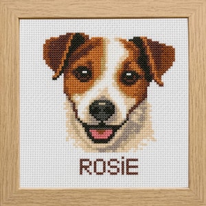 Op de afbeelding: Een kruissteekborduurwerk van een Jack Russell Terrier-hondengezicht met bruine en witte vacht, een zwarte neus en een roze tong, op een witte rasterstof. De naam ROSIE is in bruine letters onder de hond geborduurd in een lichthouten lijst.