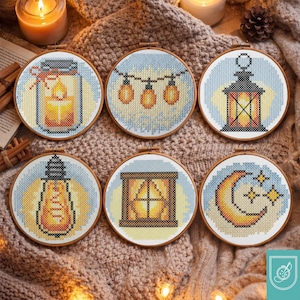 Puede incluir: Seis aros de bordado de punto de cruz redondos con varios diseños. Los diseños incluyen un tarro con una vela, luces de cadena, una linterna, una bombilla, una ventana y una luna creciente con estrellas. Cada diseño está sobre un fondo azul claro.