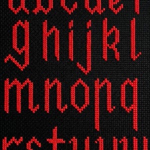 May include: Close-up of a black fabric with red cross-stitch letters of the alphabet. The letters are in a gothic font style, with each letter clearly defined against the dark background. The letters are a vibrant red.