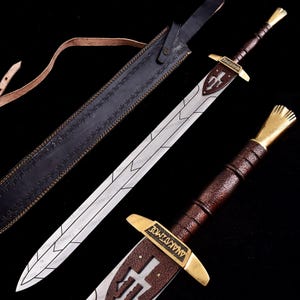 May include: A long, silver sword with a brown leather-wrapped handle and a gold-colored crossguard. The blade has etched designs and is paired with a black leather sheath with decorative stitching. The sword's crossguard has the word "ANAKAZMOE" engraved on it.