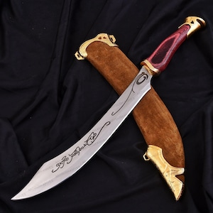 May include: A curved, silver blade knife with a red and gold handle, resting in a brown sheath with gold accents. The blade has etched designs and text. The knife is displayed on a black background.