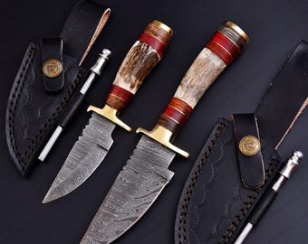 May include: Two hunting knives with Damascus steel blades, brass bolsters, and handles made of antler and red wood. The knives are displayed with black leather sheaths and sharpening steels on a dark surface.