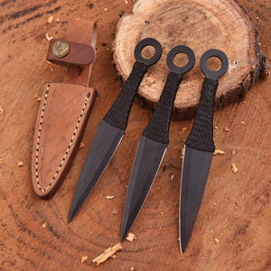 May include: Three black throwing knives with ring handles and wrapped grips are displayed on a wooden surface. A brown leather sheath with a decorative stitch pattern and a belt loop is also present. The knives have sharp, pointed blades.