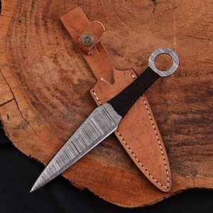 May include: A Damascus steel throwing knife with a black wrapped handle and a ring at the end. The knife is in a brown leather sheath with a snap closure. The knife and sheath are on a wooden surface.