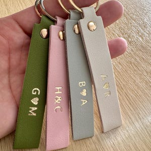 May include: Four leather keychains in olive green, pink, gray, and beige, each with gold-colored initials and a heart symbol. The keychains have gold-colored rivets and key rings.