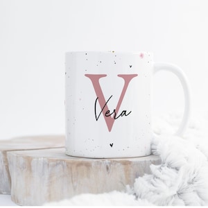 Pode incluir: Caneca de cerâmica branca com uma grande letra "V" rosa e o nome "Vera" em escrita cursiva. A caneca tem um design salpicado com pequenos corações rosa e pontos pretos. A caneca está sobre uma superfície de madeira.