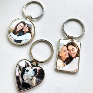 Puede incluir: Tres llaveros plateados con inserciones fotográficas. Uno es redondo, otro rectangular y otro en forma de corazón. Las fotos muestran a dos mujeres abrazándose y un perro husky con ojos azules. Los llaveros están sobre una superficie blanca.