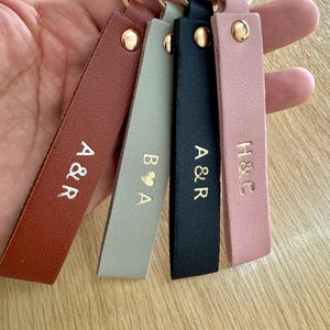 May include: A collection of personalized leather keychains in various colors, including brown, gray, black, and pink. Each keychain features gold-colored initials and a gold-tone metal ring.