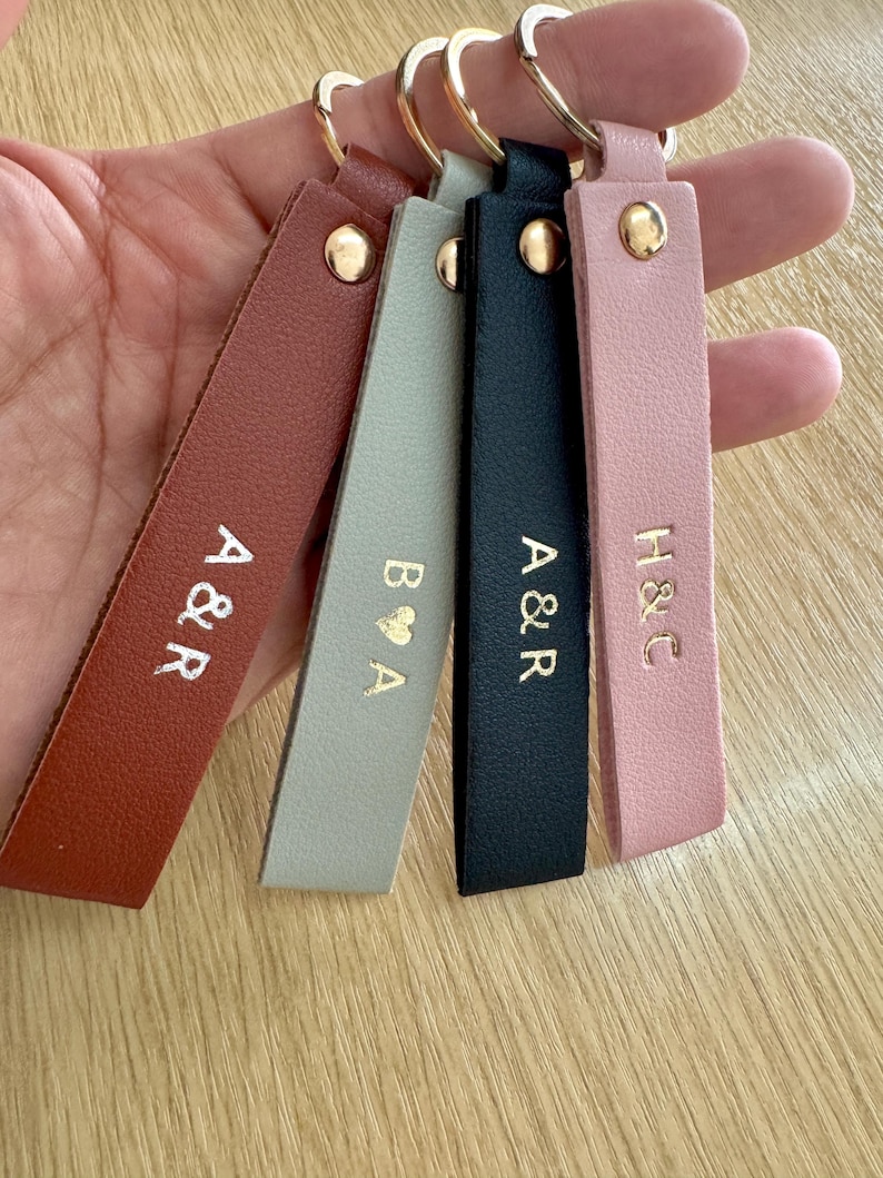 May include: A collection of personalized leather keychains in various colors, including brown, gray, black, and pink. Each keychain features gold-colored initials and a gold-tone metal ring.