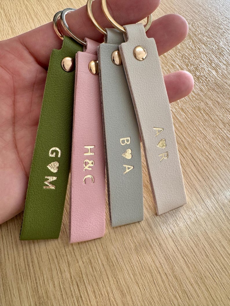 May include: Four leather keychains in olive green, pink, gray, and beige, each with gold-colored initials and a heart symbol. The keychains have gold-colored rivets and key rings.