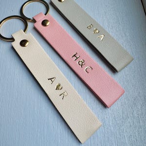 May include: Three personalized keychains in cream, pink, and gray. Each keychain features gold lettering with initials and a gold-colored metal ring. The cream keychain has "A ♡ R", the pink one has "H & C", and the gray one has "B ♡ A".