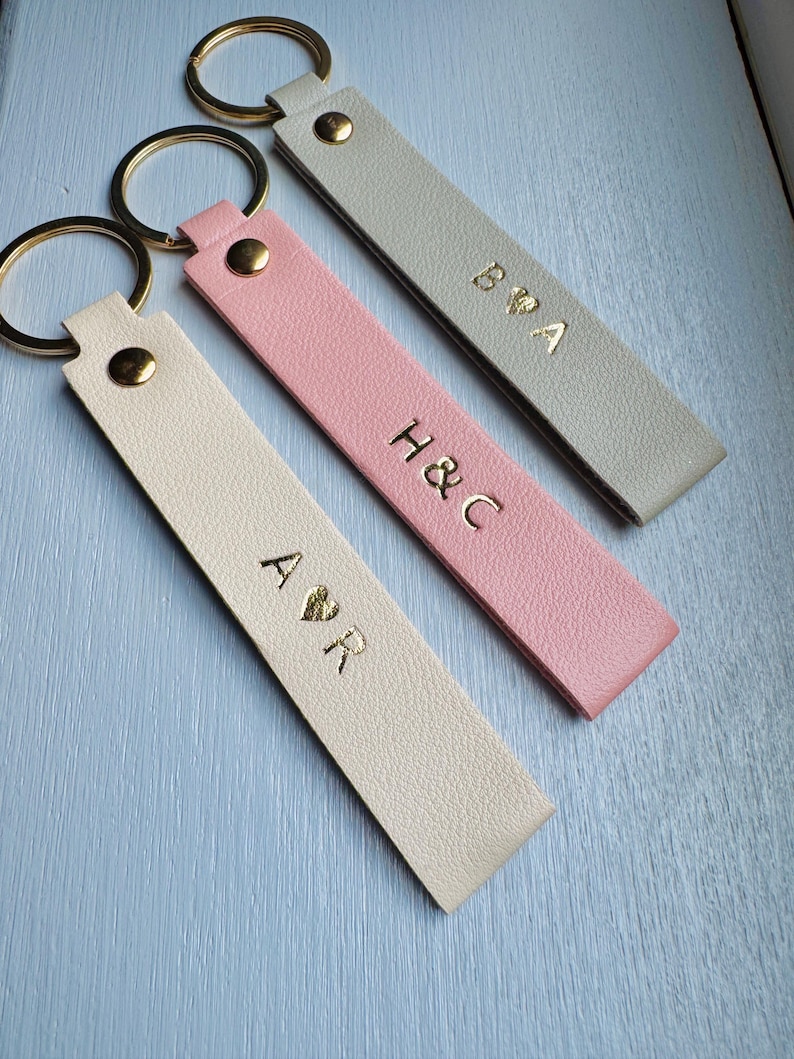 May include: Three personalized keychains in cream, pink, and gray. Each keychain features gold lettering with initials and a gold-colored metal ring. The cream keychain has "A ♡ R", the pink one has "H & C", and the gray one has "B ♡ A".