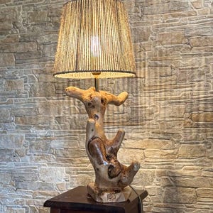 May include: A unique wooden table lamp with a natural, organic design. The lamp features a textured, light brown wooden base and a beige lampshade. The lamp is placed on a small, dark brown wooden table.