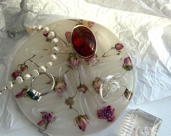 May include: A round, translucent resin tray embedded with dried pink rosebuds, featuring a large oval amber-colored cabochon, a silver ring with a green stone, two pearl necklaces, and a small square glass bottle with a clear stopper.