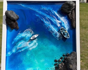 May include: A square wall art piece in a white frame featuring a resin ocean scene with shades of deep blue and turquoise. Two miniature boats float on the surface, surrounded by white resin waves, with dark textured rocks placed in the corners.