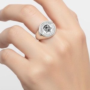 May include: A silver signet ring with a circular face featuring a detailed black and white emblem. The ring is worn on a hand against a white background. The emblem has a star-like design.