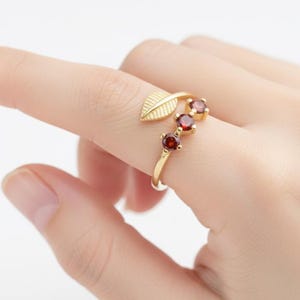 Könnte beinhalten: Ein goldfarbener Ring mit einem Blattmotiv und drei roten Edelsteinen. Der Ring wird an einem Finger getragen und zeigt die filigranen Details des Schmucks. Die Edelsteine sind in einer Gruppe angeordnet und verleihen dem Ring einen Hauch von Eleganz. Ein Schmuckstück.