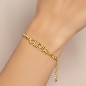 Puede incluir: Una pulsera dorada con el nombre "Olivia" en letras mayúsculas. La pulsera está en una cadena delicada y se lleva en la muñeca. El fondo es de un color suave y neutro.