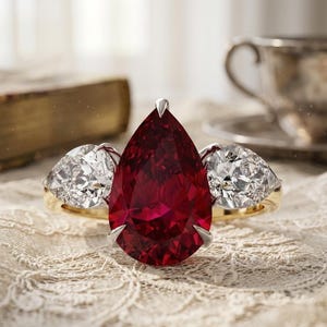 Puede incluir: Un anillo de oro con una gran piedra preciosa roja en forma de pera como pieza central, flanqueada por dos piedras blancas más pequeñas, talladas en forma triangular. El anillo está sobre una superficie de encaje.