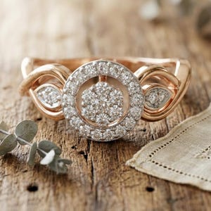 Puede incluir: Anillo de oro rosa con un grupo central de diamantes rodeado por un halo de diamantes más pequeños. El anillo presenta un diseño único con detalles de diamantes en los laterales, sobre una superficie de madera rústica.
