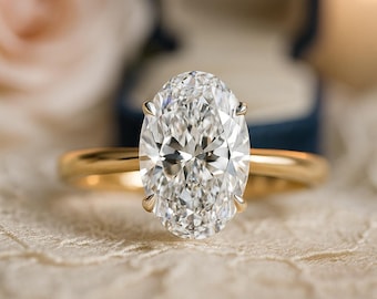 May include: A gold engagement ring with a large, oval-cut diamond. The ring is set on a delicate, lace-like surface, with a blurred background including a blue ring box and a soft, pink flower.