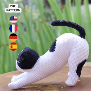May include: A crochet cat toy featuring a white body with black patches on the head, back, and hind leg. The cat is in a stretching pose with its front paws extended forward and its tail curved upward. The image includes a PDF PATTERN label.