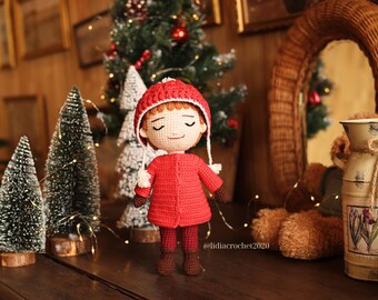 Puede incluir: Muñeca de ganchillo hecha a mano con abrigo, gorro y pantalón rojos, con botas y guantes marrones. La muñeca está de pie frente a un pequeño árbol de Navidad y otras decoraciones navideñas. La muñeca mide aproximadamente 15 cm.
