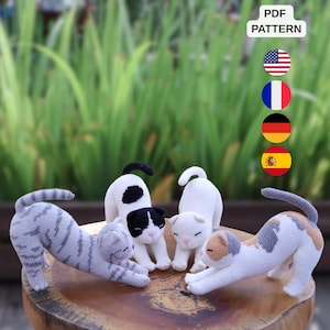 May include: Four crocheted cat figurines in a stretching pose. One is grey tabby, one is black and white, one is solid white, and one is calico with white, grey, and tan patches. Each cat has embroidered eyes and a pink nose. Text reads PDF PATTERN.