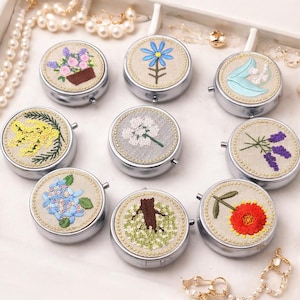 May include: A collection of nine small, round, silver-colored pill boxes. Each box features a different embroidered floral design on the lid, including lavender, blue flowers, and a red gerbera daisy. The boxes are arranged on a white surface.