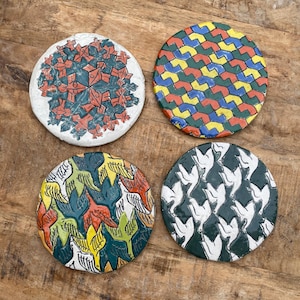 May include: Four round ceramic coasters with colorful, geometric designs. One coaster features a red and green floral pattern, another has a tessellated pattern in red, yellow, green, and blue. The other two coasters have bird and dove patterns.