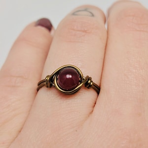 May include: A simple wire-wrapped ring with a dark red gemstone. The ring is made of brass wire and features a single, round gemstone.