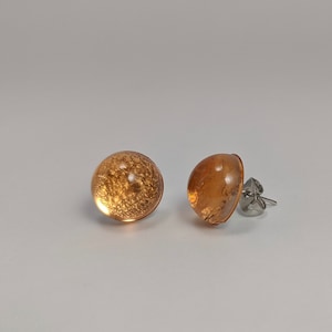 May include: A pair of gold-toned stud earrings with a round, clear glass design. The glass has a sparkly, glittery effect.