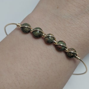 May include: A gold wire bangle bracelet with five green, brown, and gold speckled round beads.