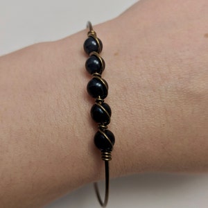 May include: A simple bracelet with a gold wire base and five black beads. The beads are wrapped in wire and are evenly spaced along the bracelet.