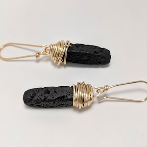 May include: A pair of black lava rock earrings with gold wire wrapped around the stone. The earrings have a simple hook closure.