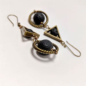 May include: A pair of black lava rock earrings with gold geometric wire accents. The earrings feature a circle, a triangle, and a small round bead.