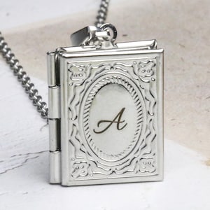Puede incluir: Colgante plateado en forma de libro con cadena. El medallón presenta un diseño ovalado con la letra "A" en el centro. El colgante está detallado con patrones decorativos y bisagras.