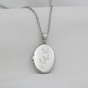 Puede incluir: Collar con colgante ovalado plateado y cadena fina. El medallón presenta un diseño grabado de tres mariposas. El collar se muestra sobre un fondo blanco.