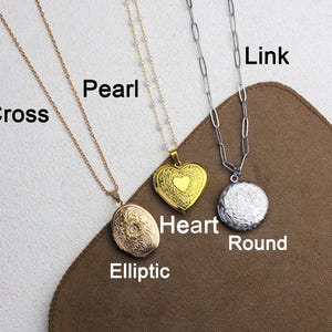May include: Three necklaces with pendants are displayed. One is gold with an oval locket, another is gold with a heart-shaped locket, and the third is silver with a round pendant. The necklaces are on a brown surface.