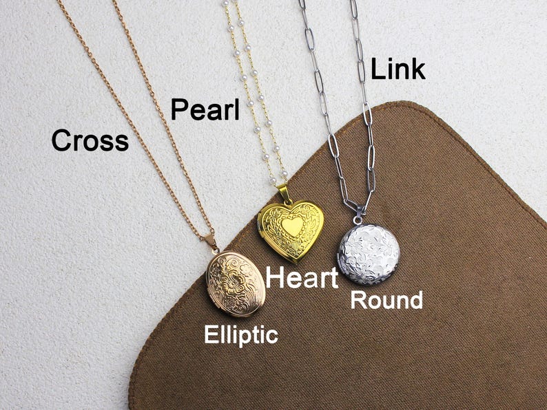 May include: Three necklaces with pendants are displayed. One is gold with an oval locket, another is gold with a heart-shaped locket, and the third is silver with a round pendant. The necklaces are on a brown surface.