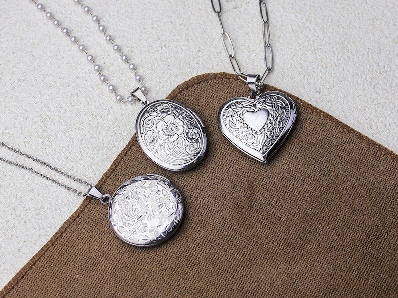May include: Three silver lockets with intricate floral and heart designs. One oval, one round, and one heart-shaped locket, each suspended from a delicate chain. The lockets rest on a brown fabric surface, showcasing their detailed craftsmanship.