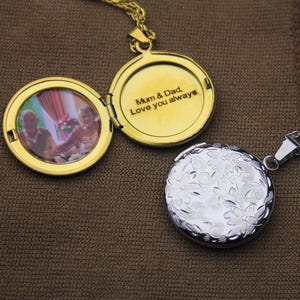 May include: Two open gold-colored lockets and one closed silver-colored locket on a brown surface. The open locket displays a photo of an older couple and the inscription "Mum & Dad, Love you always."