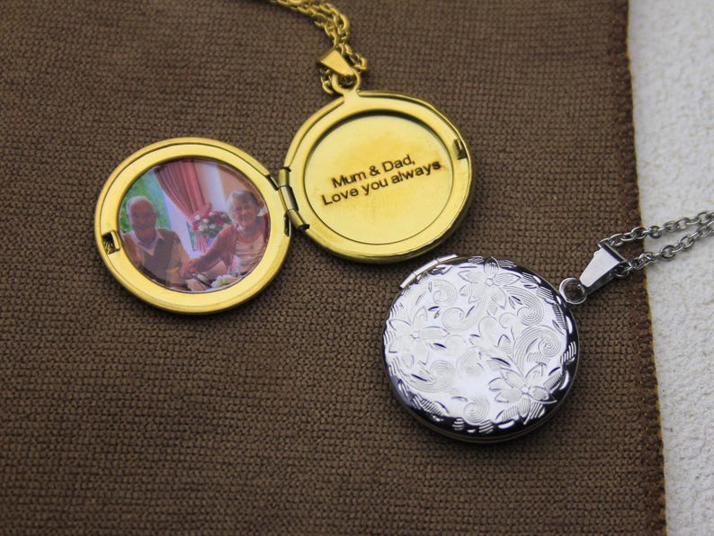 May include: Two open gold-colored lockets and one closed silver-colored locket on a brown surface. The open locket displays a photo of an older couple and the inscription "Mum & Dad, Love you always."