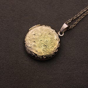 May include: A gold-colored, round locket pendant with a floral design, suspended from a silver-colored chain. The locket is intricately detailed with embossed flowers and swirling patterns. The background is a dark gray fabric.