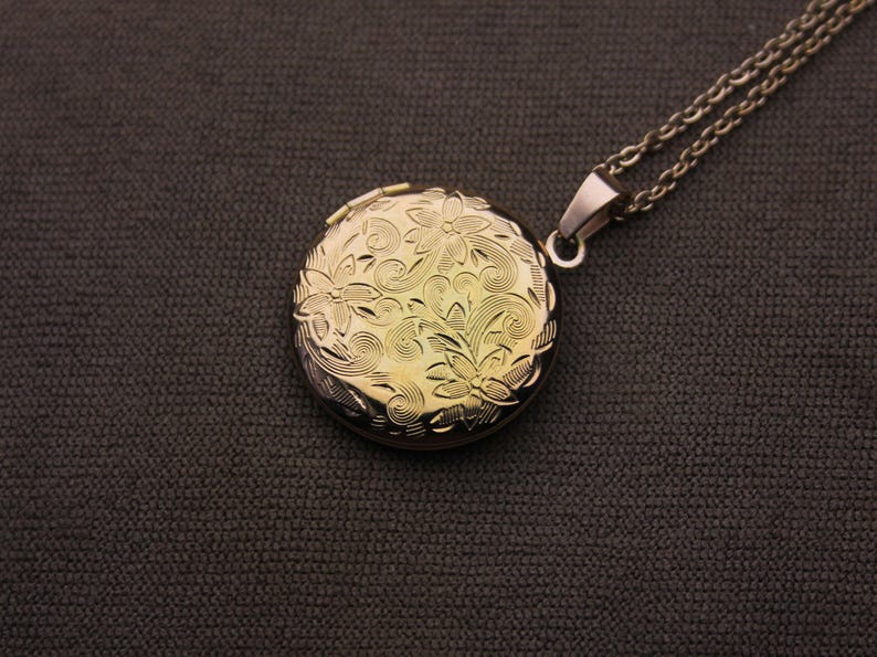 May include: A gold-colored, round locket pendant with a floral design, suspended from a silver-colored chain. The locket is intricately detailed with embossed flowers and swirling patterns. The background is a dark gray fabric.