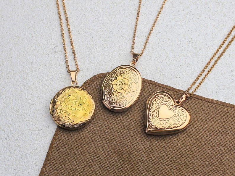 May include: Three gold-colored lockets with delicate floral and heart designs. Each locket hangs from a matching chain. The lockets are round, oval, and heart-shaped, resting on a brown fabric.