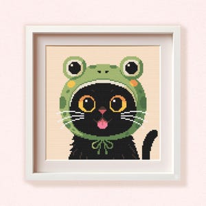 May include: A framed cross-stitch art piece featuring a black cat wearing a green frog hat. The cat has large yellow eyes, white whiskers, and a pink tongue sticking out. The artwork is set against a beige background.