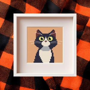 Peut inclure: Point de croix encadré d'un chat avec une fourrure noire et blanche, des yeux jaunes et des moustaches blanches. Le chat est centré sur un fond beige dans un cadre blanc. Le fond est un tissu à carreaux noir et orange.
