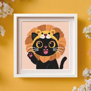 May include: A framed cross-stitch of a black cat wearing a lion costume, waving with its paw. The cat has large yellow eyes and a pink mouth. The lion's mane and hat are shades of brown and tan. The background is a light peach color.